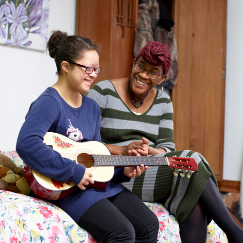 Two people sitting on a bed playing a guitar and laughing